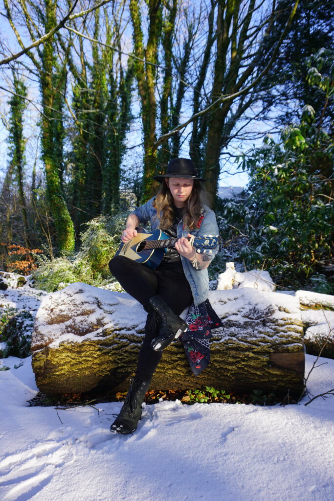 Photo of Northern Irish Blues Rock Artist Callum Norris holding a guitar sitting on a snowy log